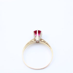 May include: A gold ring with a red gemstone set in a white metal setting. The ring has a delicate, ornate design.