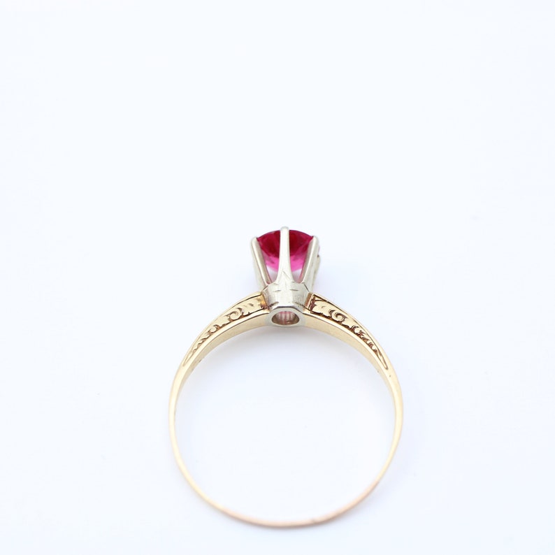 May include: A gold ring with a red gemstone set in a white metal setting. The ring has a delicate, ornate design.