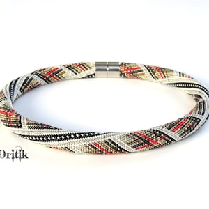 May include: A white, black, gold, and red beaded necklace with a magnetic clasp. The necklace is made of small beads in a geometric pattern.