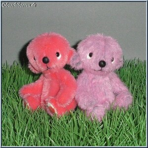 Puede incluir: Dos figuras de osos de peluche, una roja y otra lavanda, sentadas en hierba verde artificial. Los osos tienen narices de botón negras y ojos con borde blanco. Los osos están sentados erguidos, con los brazos y las piernas visibles.