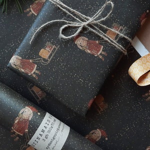 May include: Black and white wrapping paper with a repeating pattern of a girl pulling a sled with a gift on it. The paper has a subtle sparkle effect. The text "FINA MAIS" is printed on the paper.