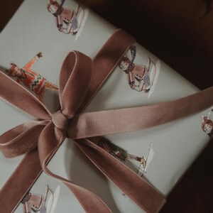 May include: A wrapped gift with a light blue background featuring illustrations of people skiing. The gift is tied with a dusty rose velvet ribbon and bow. The overall theme is winter and gift-giving.