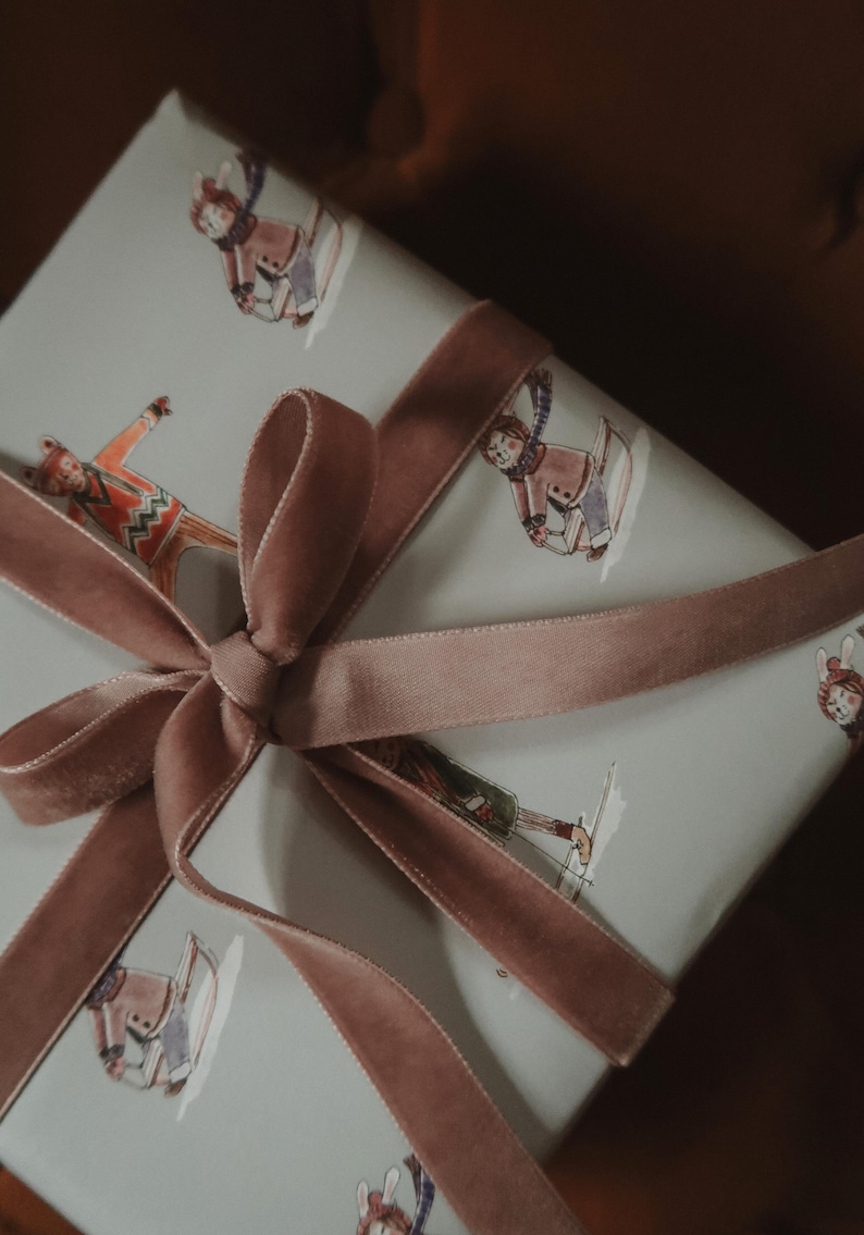 May include: A wrapped gift with a light blue background featuring illustrations of people skiing. The gift is tied with a dusty rose velvet ribbon and bow. The overall theme is winter and gift-giving.