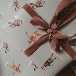 May include: A wrapped gift with light gray paper featuring whimsical skier illustrations in various outfits. A dusty rose velvet ribbon is tied in a bow. The gift is ready for the holidays.