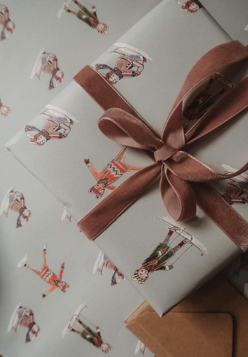 May include: A wrapped gift with light gray paper featuring whimsical skier illustrations in various outfits. A dusty rose velvet ribbon is tied in a bow. The gift is ready for the holidays.