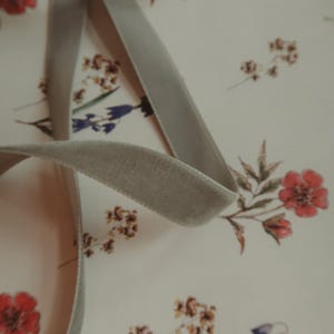 May include: A close-up of a light gray velvet ribbon on a white background with a floral pattern. The ribbon is folded and twisted, showing its texture.