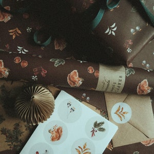 May include: A sheet of white paper with nine round stickers featuring illustrations of squirrels, leaves, and other woodland elements. The stickers are arranged in a grid pattern. The paper is lying on top of brown wrapping paper with a squirrel design. The text "WRAPPING 3 pieces mydesign" is visible on the wrapping paper.