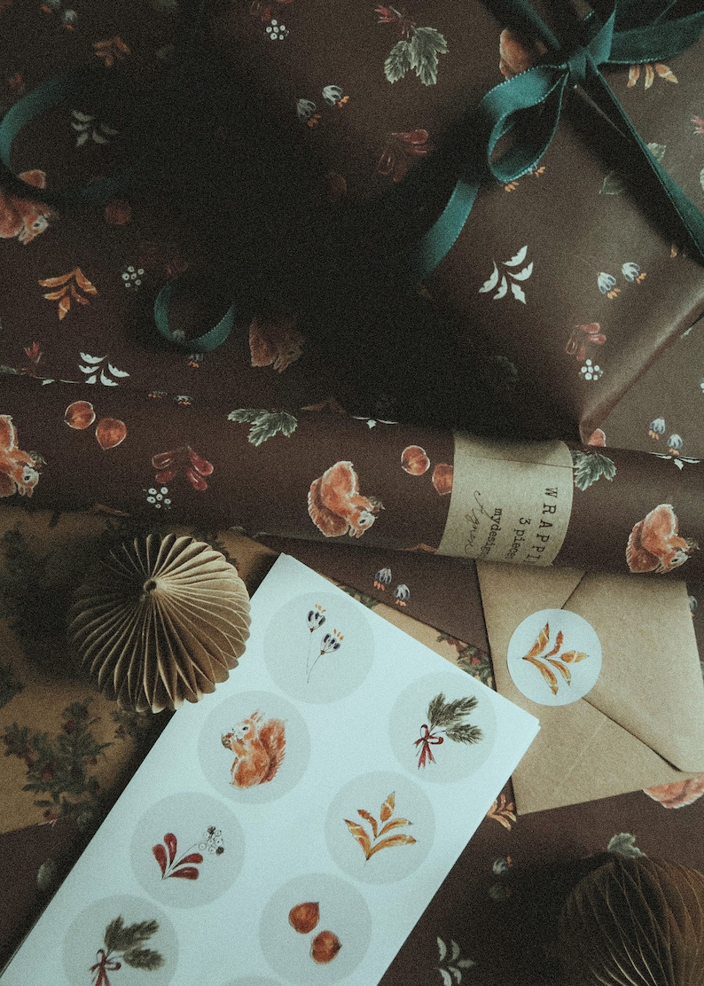May include: A sheet of white paper with nine round stickers featuring illustrations of squirrels, leaves, and other woodland elements. The stickers are arranged in a grid pattern. The paper is lying on top of brown wrapping paper with a squirrel design. The text "WRAPPING 3 pieces mydesign" is visible on the wrapping paper.
