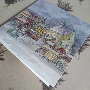 May include: A watercolor painting of a European Christmas market scene with a tall building in the background and several market stalls in the foreground. The painting is done in shades of brown, yellow, and blue.