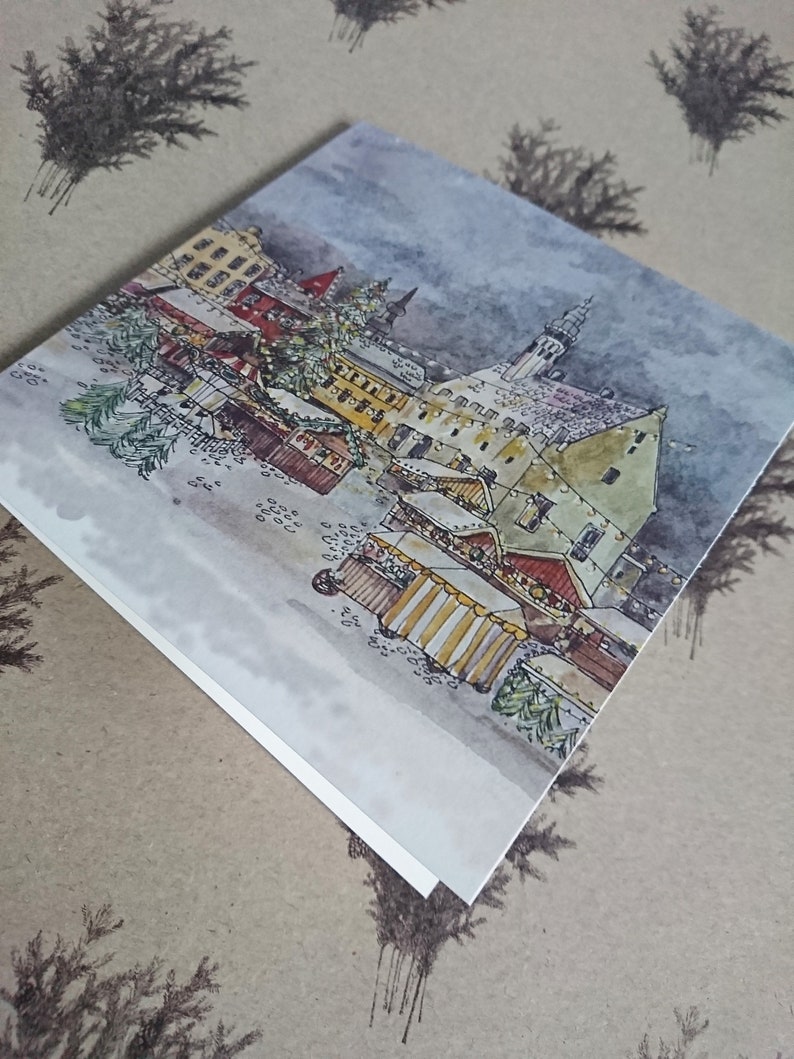 May include: A watercolor painting of a European Christmas market scene with a tall building in the background and several market stalls in the foreground. The painting is done in shades of brown, yellow, and blue.