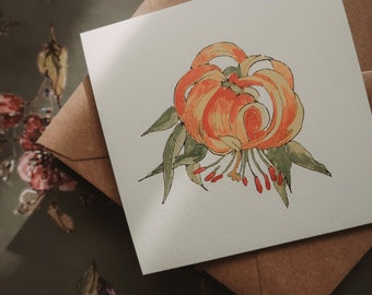 May include: A watercolor painting of an orange lily flower with green leaves on a white card. The card is on a brown envelope.