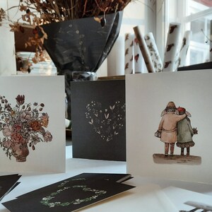 May include: Four greeting cards with different designs. One card features a floral bouquet in a vase, another has a heart-shaped design with small flowers, and the third card shows two people walking together. The fourth card is blank.