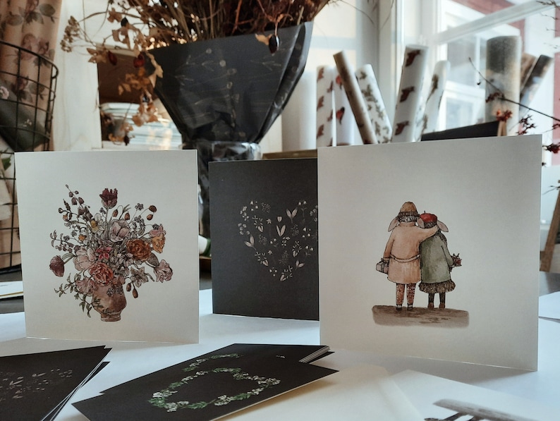May include: Four greeting cards with different designs. One card features a floral bouquet in a vase, another has a heart-shaped design with small flowers, and the third card shows two people walking together. The fourth card is blank.