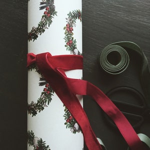 May include: A white wrapping paper roll with a repeating pattern of green wreaths with red berries. A red velvet ribbon is tied around the roll.