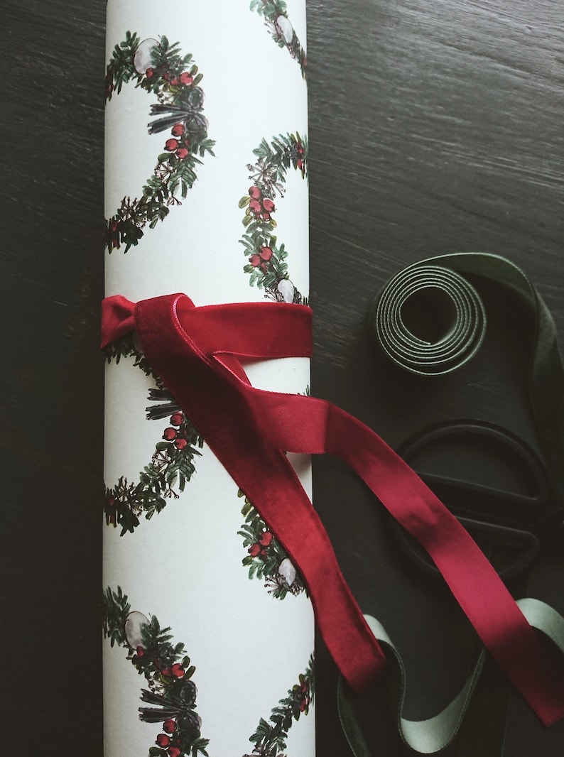 May include: A white wrapping paper roll with a repeating pattern of green wreaths with red berries. A red velvet ribbon is tied around the roll.
