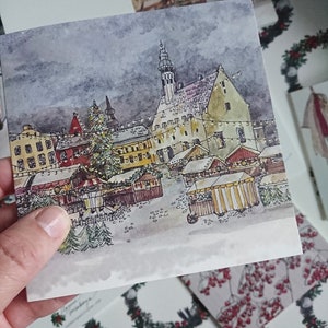 May include: A watercolor painting of a European Christmas market scene with a tall church steeple in the background. The market is filled with wooden stalls and decorated with twinkling lights. There is a large Christmas tree in the center of the market.