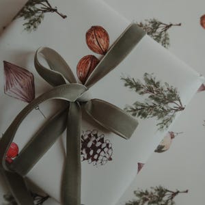 May include: A white gift box with a green velvet ribbon tied in a bow. The box is wrapped in paper with a repeating pattern of pine branches, acorns, and berries.
