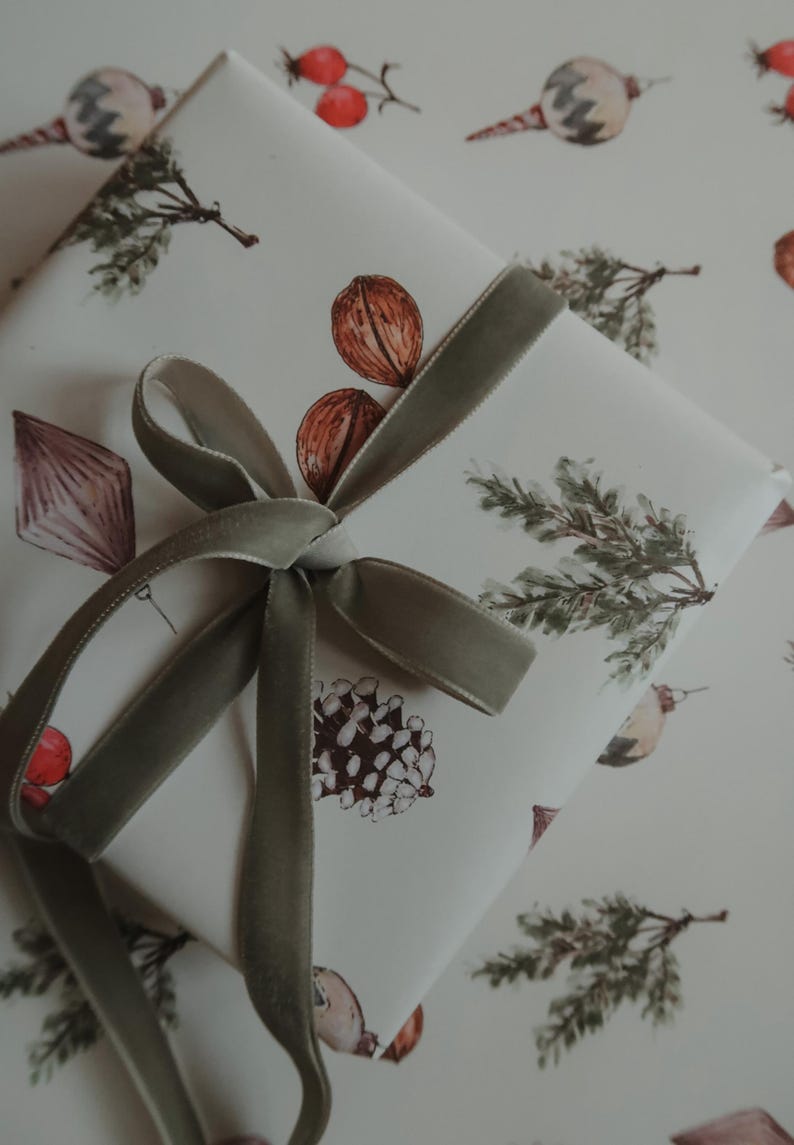 May include: A white gift box with a green velvet ribbon tied in a bow. The box is wrapped in paper with a repeating pattern of pine branches, acorns, and berries.