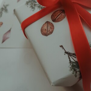May include: A white gift box with a red ribbon tied in a bow. The box is decorated with a simple design of nuts and leaves.