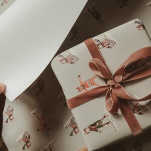 May include: A wrapped gift with a brown velvet ribbon and bow, and matching wrapping paper. The paper has a light gray background with illustrations of skiers in various outfits. A hand is holding a sheet of the wrapping paper.
