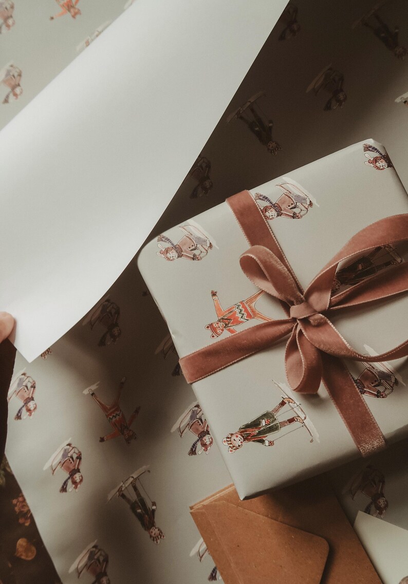 May include: A wrapped gift with a brown velvet ribbon and bow, and matching wrapping paper. The paper has a light gray background with illustrations of skiers in various outfits. A hand is holding a sheet of the wrapping paper.