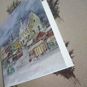 May include: A watercolor painting of a snowy European village with a Christmas market. The painting features a tall building with a pointed roof, a red-roofed building, and several market stalls with striped awnings. The painting is done in a muted color palette with shades of gray, brown, and white.