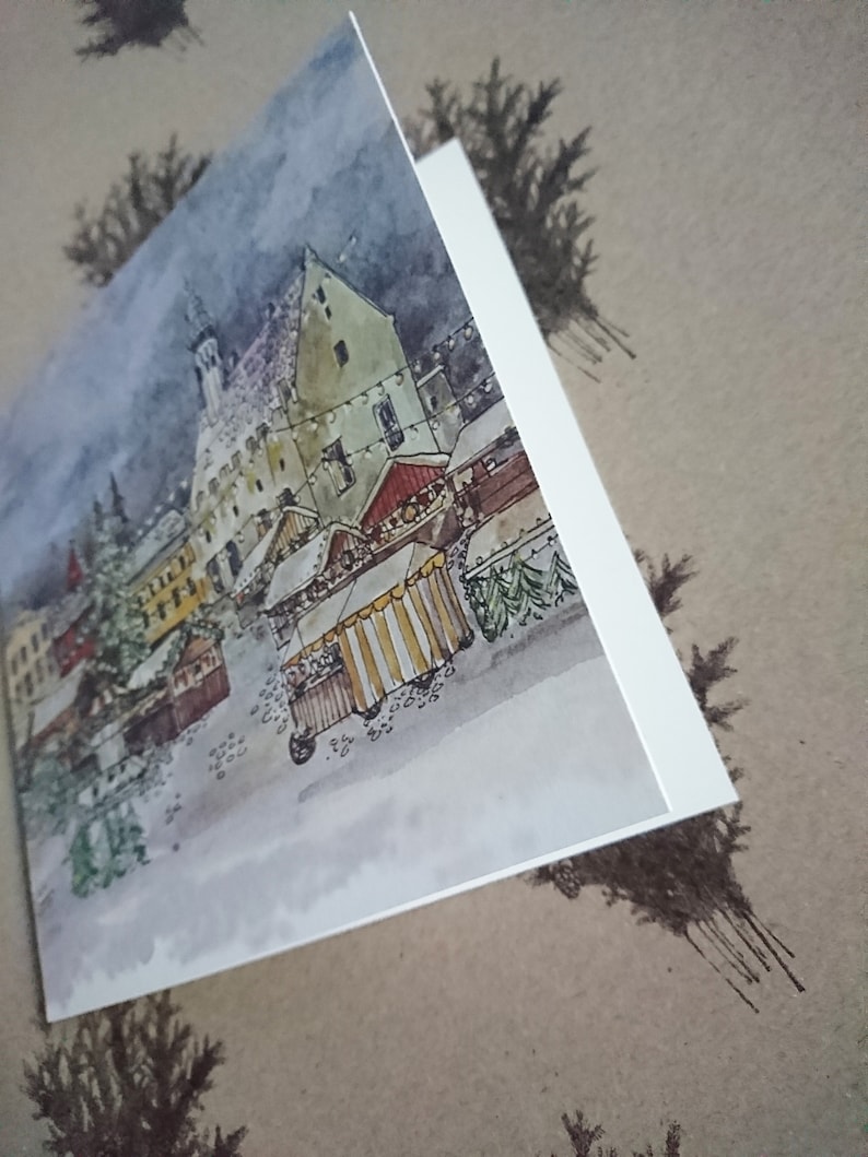 May include: A watercolor painting of a snowy European village with a Christmas market. The painting features a tall building with a pointed roof, a red-roofed building, and several market stalls with striped awnings. The painting is done in a muted color palette with shades of gray, brown, and white.
