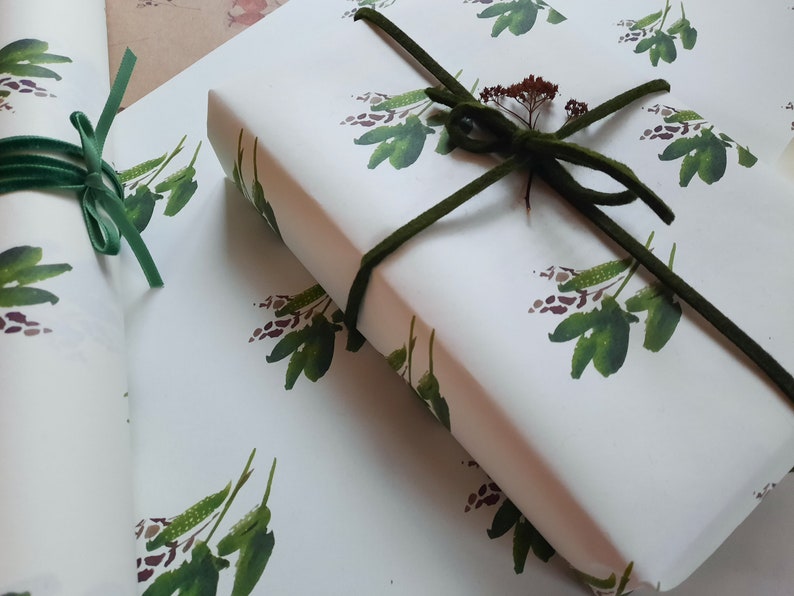 May include: Two rolls of white wrapping paper with a green floral pattern. The paper is tied with a green ribbon and a small sprig of dried flowers.