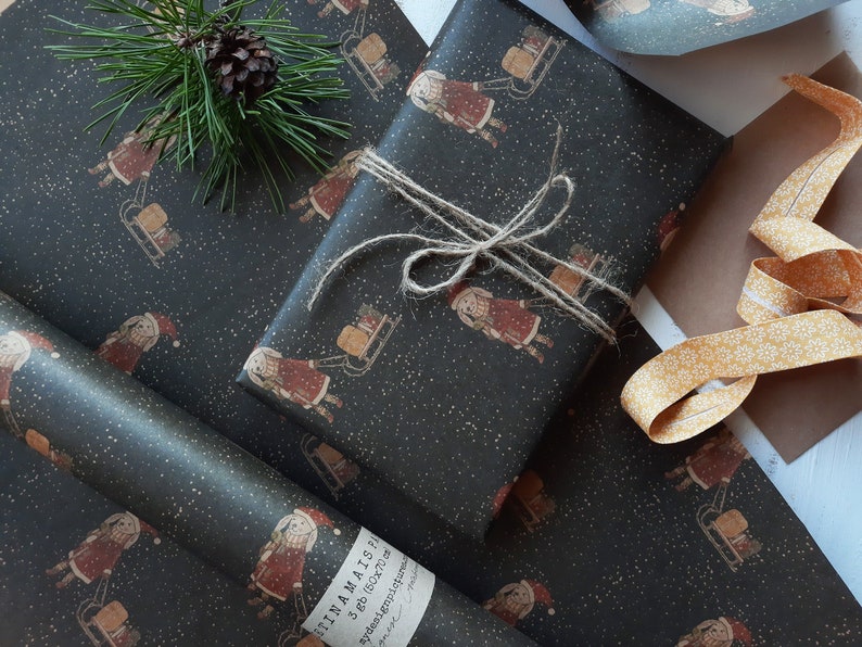 May include: Black and white wrapping paper with a repeating pattern of a girl pulling a sled with a gift on it. The paper has a subtle sparkle effect. The text "FINA MAIS" is printed on the paper.