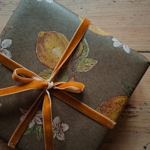 Pode incluir: Uma caixa de presente retangular embrulhada em papel verde-oliva escuro com um padrão repetitivo de limões amarelos, folhas verdes e flores brancas. O pacote está amarrado com uma fita de veludo amarelo-mostarda vibrante em um laço.