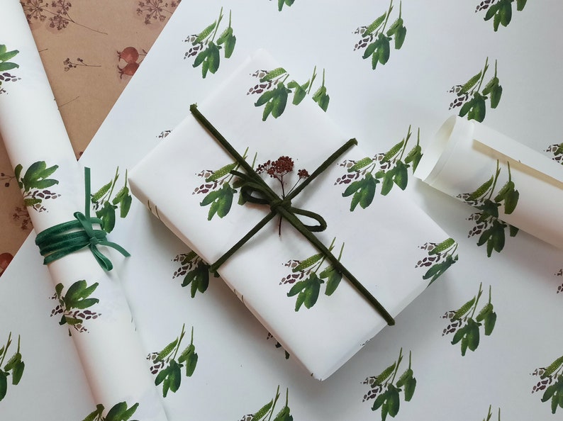 May include: White wrapping paper with a repeating pattern of green leafy sprigs. Two rolls of the paper are shown, one is wrapped around a gift box with a green ribbon.