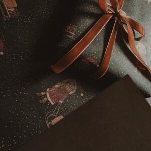 May include: A brown gift box wrapped in dark green paper with a white snowflake pattern. The box is tied with a brown satin ribbon with a bow.