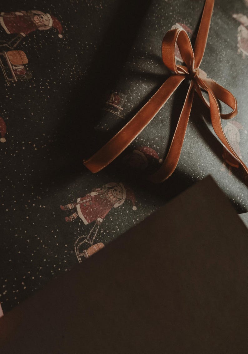 May include: A brown gift box wrapped in dark green paper with a white snowflake pattern. The box is tied with a brown satin ribbon with a bow.