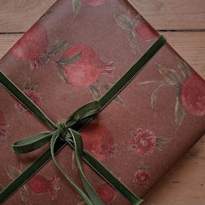 Könnte beinhalten: Ein rechteckiges Geschenk, eingewickelt in braunes Papier mit einem sich wiederholenden Muster aus roten Granatäpfeln und grünen Blättern. Das Paket ist mit einem dünnen, dunkelgrünen Samtband umwickelt, das vorne zu einer Schleife gebunden ist.