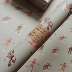 May include: A roll of gray wrapping paper with a repeating pattern of skiers in winter attire. A wrapped gift box with a brown ribbon sits in the background. The paper roll has a label that reads "Wrapping Paper (50x70 cm)."