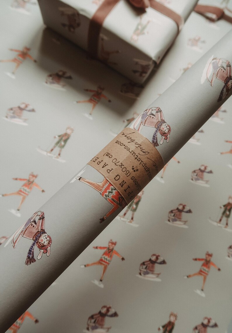 May include: A roll of gray wrapping paper with a repeating pattern of skiers in winter attire. A wrapped gift box with a brown ribbon sits in the background. The paper roll has a label that reads "Wrapping Paper (50x70 cm)."
