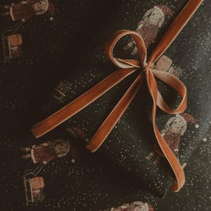 May include: A brown velvet ribbon tied in a bow on top of dark green wrapping paper with a repeating pattern of a person in a red and white outfit riding a sled. The paper is covered in white dots.