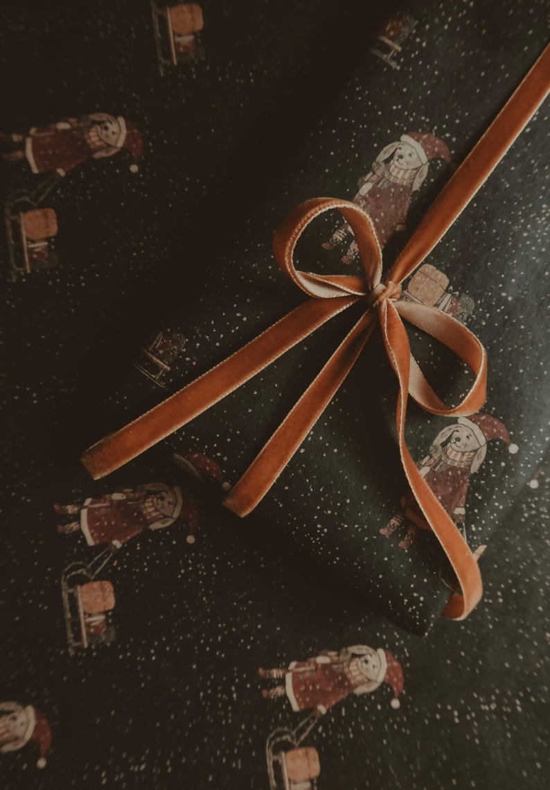 May include: A brown velvet ribbon tied in a bow on top of dark green wrapping paper with a repeating pattern of a person in a red and white outfit riding a sled. The paper is covered in white dots.