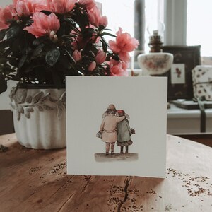 May include: A white card with a watercolor illustration of two people walking hand-in-hand. The people are wearing winter clothing and are facing away from the viewer. The card is sitting on a wooden table.
