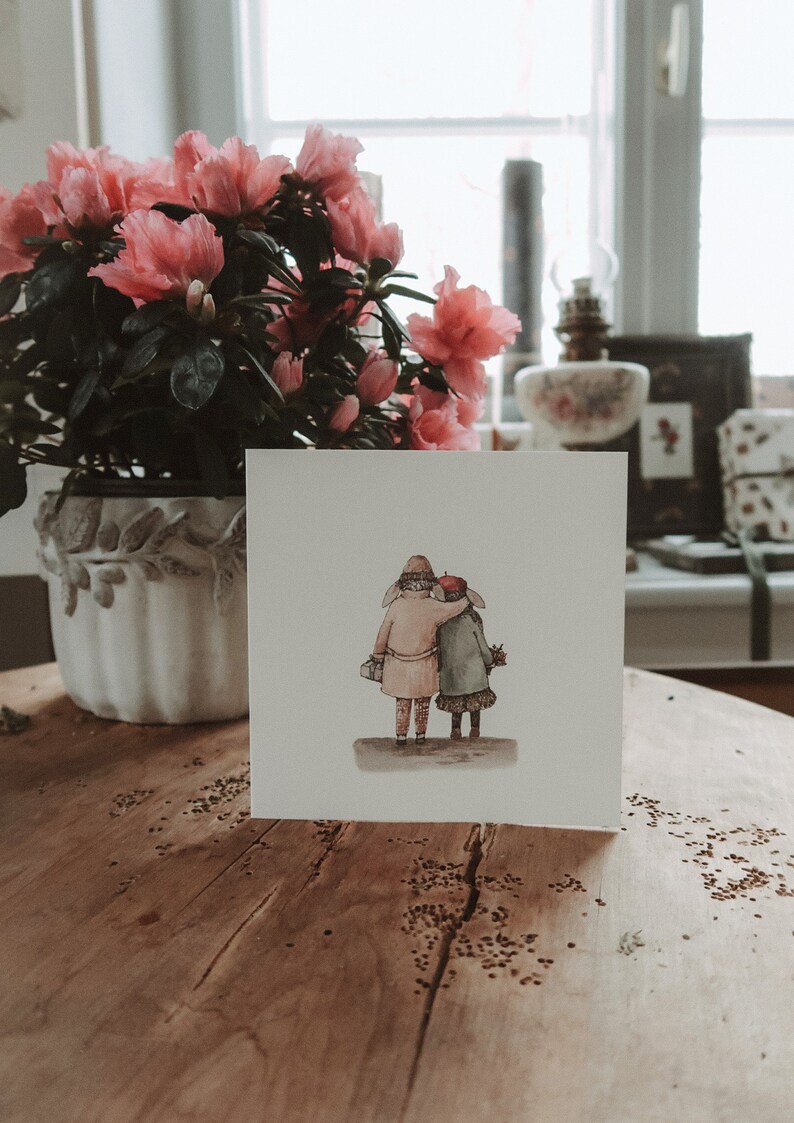 May include: A white card with a watercolor illustration of two people walking hand-in-hand. The people are wearing winter clothing and are facing away from the viewer. The card is sitting on a wooden table.