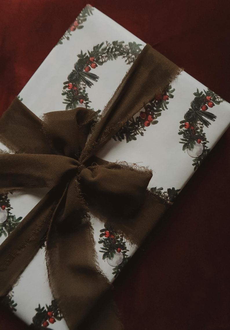 May include: A white gift box with a brown ribbon tied in a bow. The box is wrapped in paper with a repeating pattern of green wreaths with red berries.