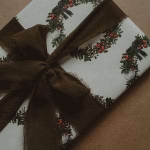 May include: A white gift box with a brown ribbon tied in a bow. The box is wrapped in paper with a repeating pattern of green wreaths with red berries.