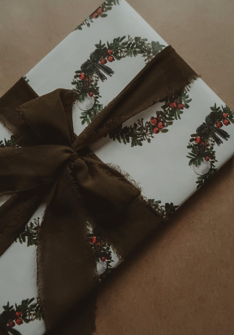 May include: A white gift box with a brown ribbon tied in a bow. The box is wrapped in paper with a repeating pattern of green wreaths with red berries.