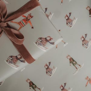 May include: Gift wrap featuring a repeating pattern of skiers in winter attire on a light gray background. A brown velvet ribbon is tied around a wrapped gift. The skiers are in various poses, wearing hats, scarves, and coats.