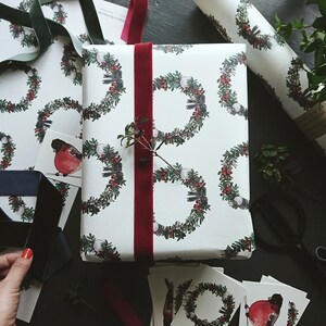 May include: A white gift wrap with a repeating pattern of green wreaths with red berries and white snowballs. A red robin bird is featured on one of the wreaths. The gift is wrapped with a red velvet ribbon.