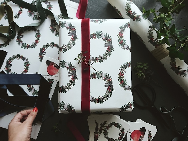 May include: A white gift wrap with a repeating pattern of green wreaths with red berries and white snowballs. A red robin bird is featured on one of the wreaths. The gift is wrapped with a red velvet ribbon.