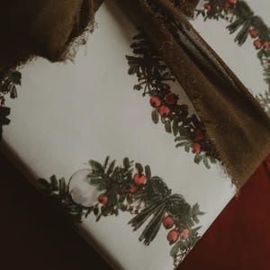 May include: A white gift wrap with a repeating pattern of green wreaths with red berries. A brown ribbon is tied around the gift.