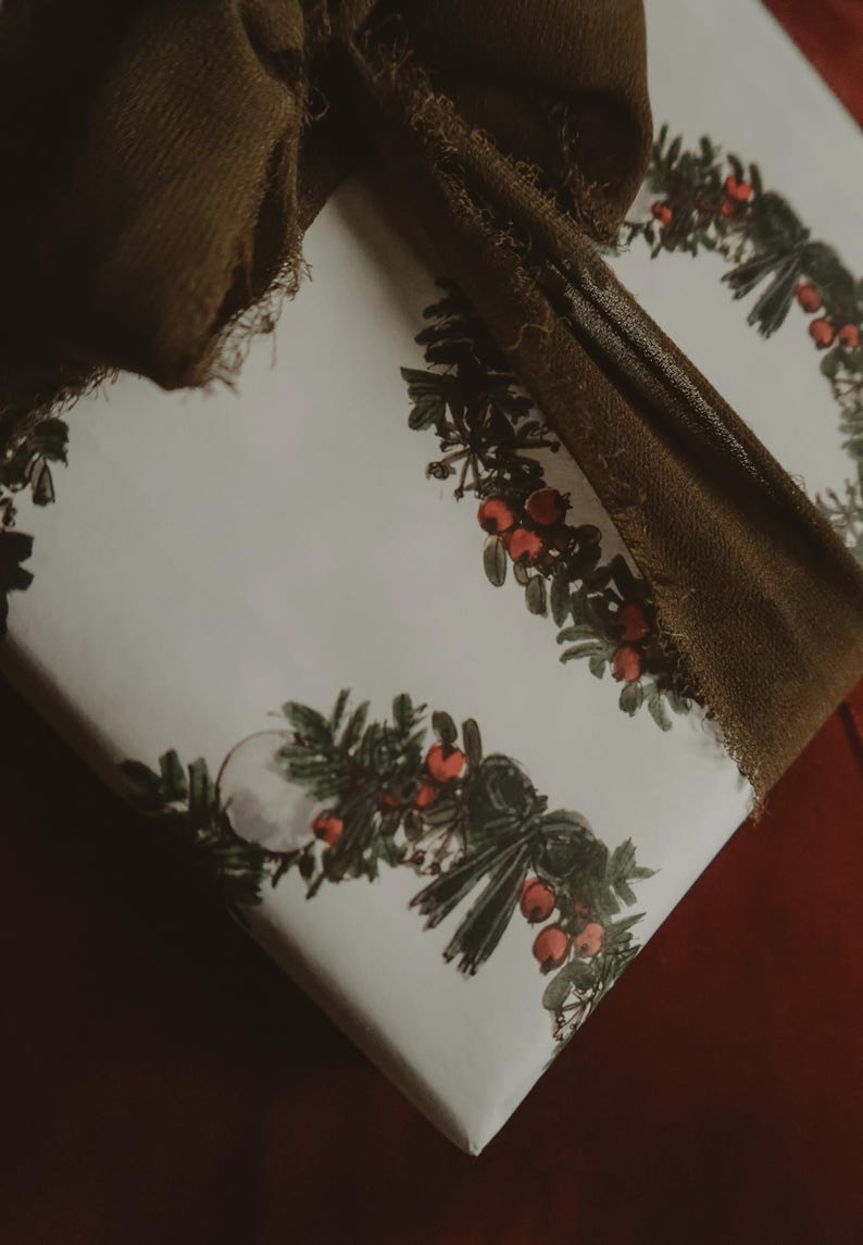 May include: A white gift wrap with a repeating pattern of green wreaths with red berries. A brown ribbon is tied around the gift.