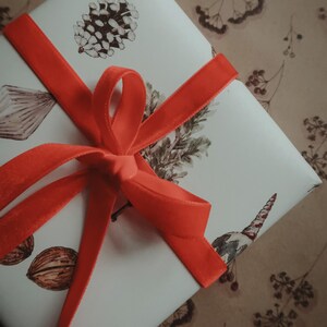 May include: A white gift box with a red velvet ribbon tied in a bow. The box is decorated with a pine cone, a sprig of greenery, and other botanical illustrations.