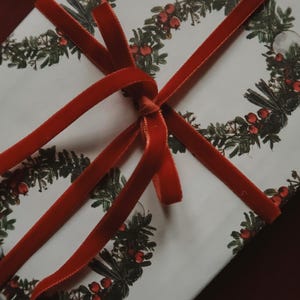 May include: A white gift box with a red velvet ribbon tied in a bow. The box is wrapped in paper with a repeating pattern of green wreaths with red berries.
